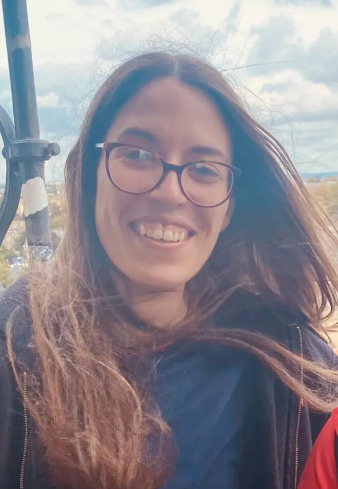 A photo of Laurie Doyle, a person with long brown hair and glasses, wearing a blue t-shirt and black hoodie. They are smiling at the camera with a daytime cityscape behind them.
